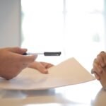Side view of faceless formal man giving pen and paper to focused female with clenched hands at table on meeting
