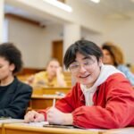 Cheerful student wearing glasses seated in a university lecture hall takes notes during a class.