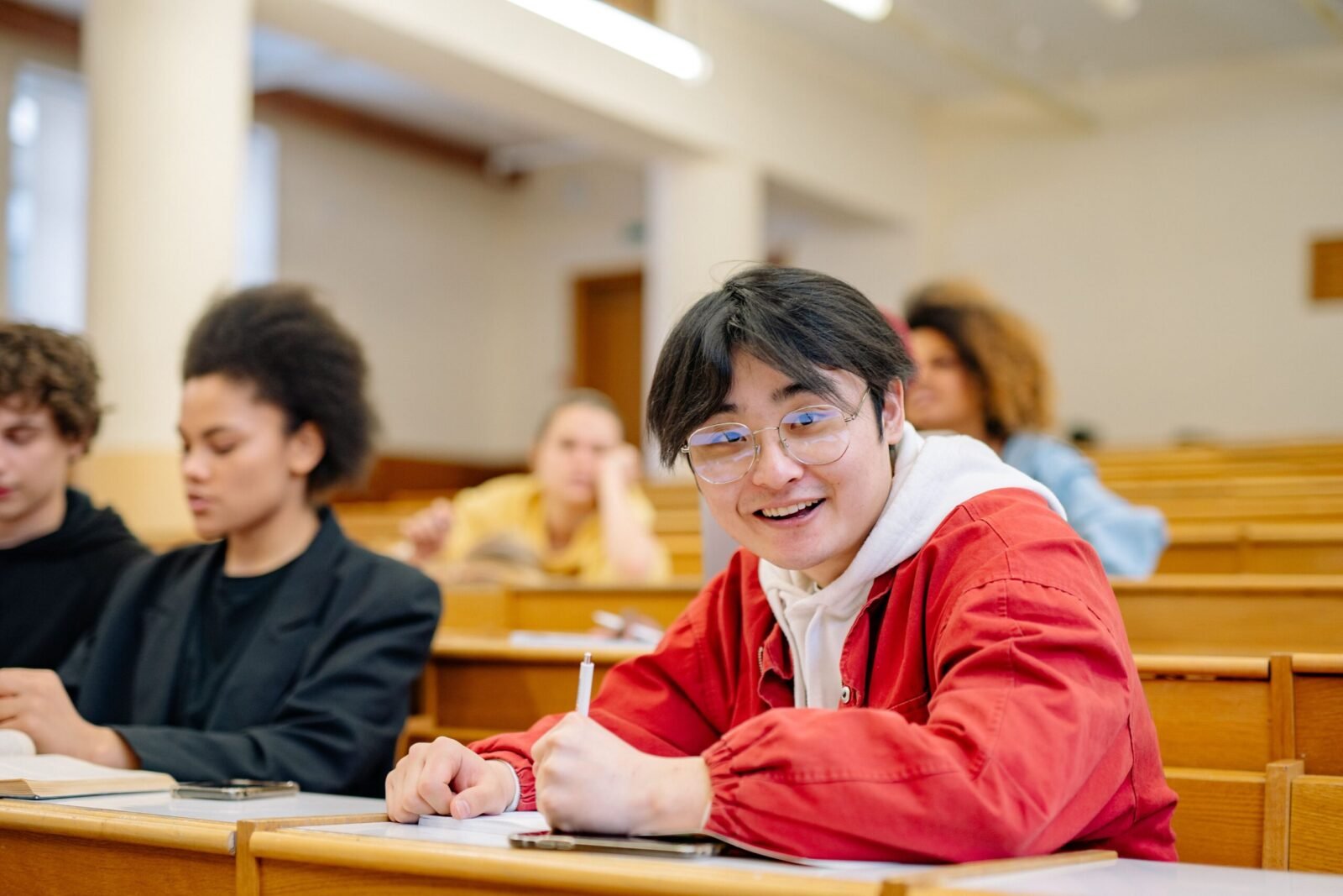 Cheerful student wearing glasses seated in a university lecture hall takes notes during a class.